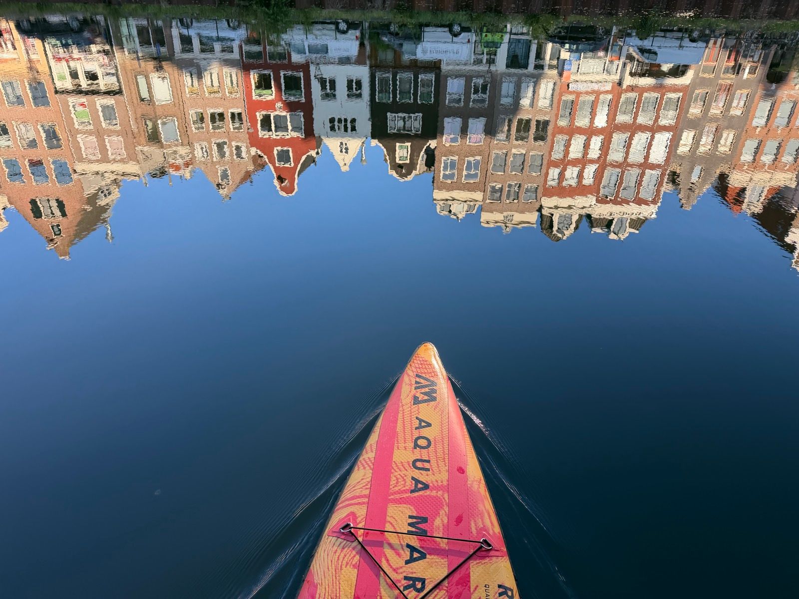 The image captures a vibrant orange and yellow paddleboard gliding across a calm, deep blue canal
