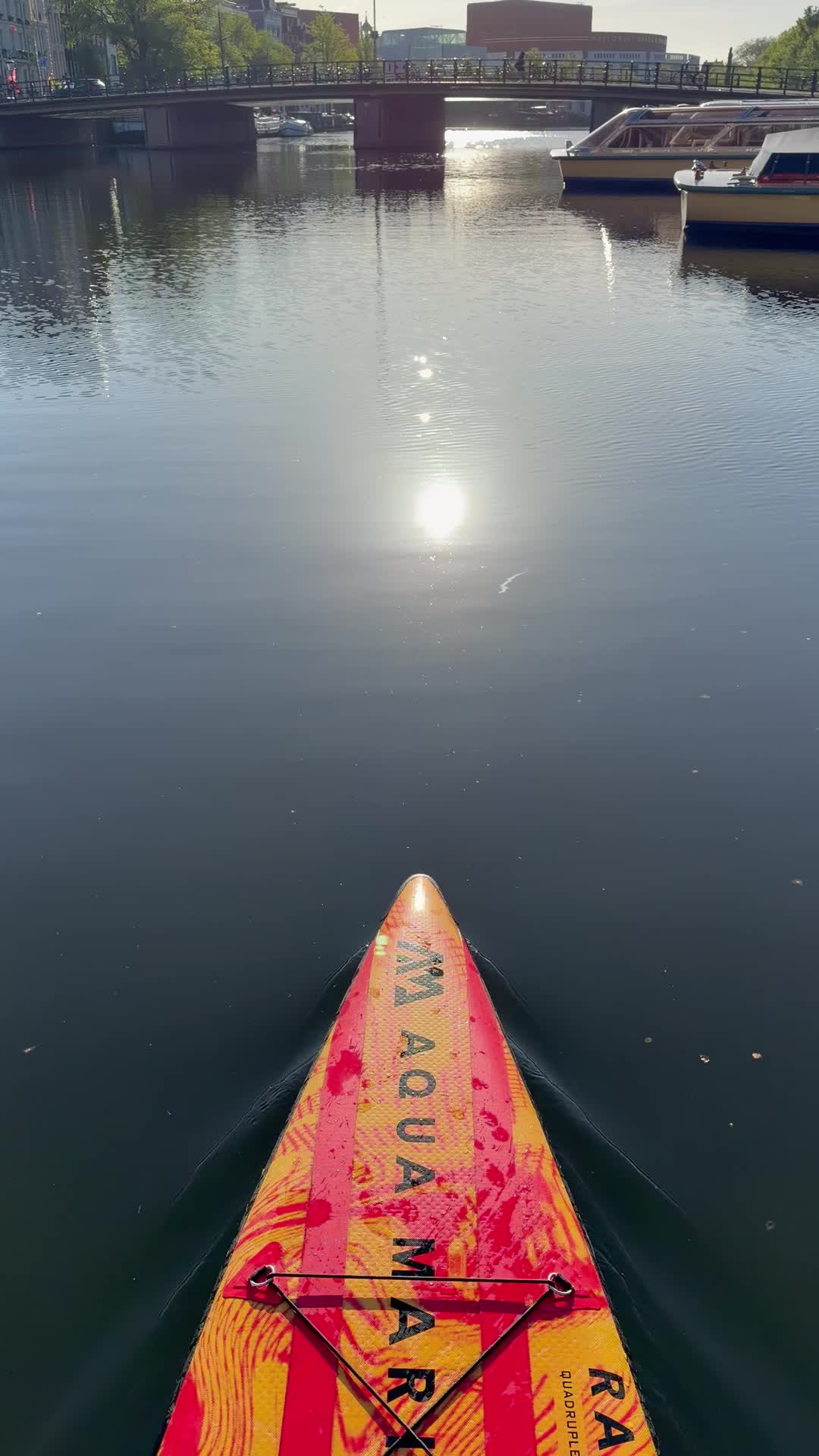 The photo captures a first-person view from a vibrant red and yellow paddleboard gliding across calm water, with the bright morning sun reflecting intensely on the surface