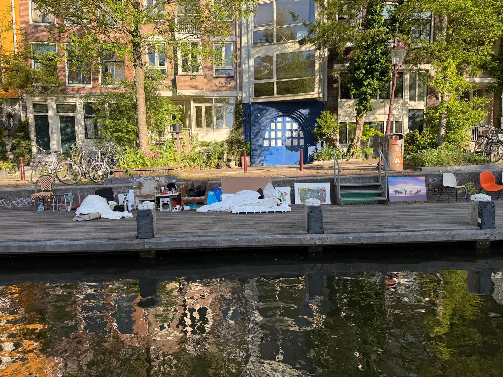 This photo captures a serene canal scene with two individuals sleeping on mattresses on a wooden dock, surrounded by various personal belongings