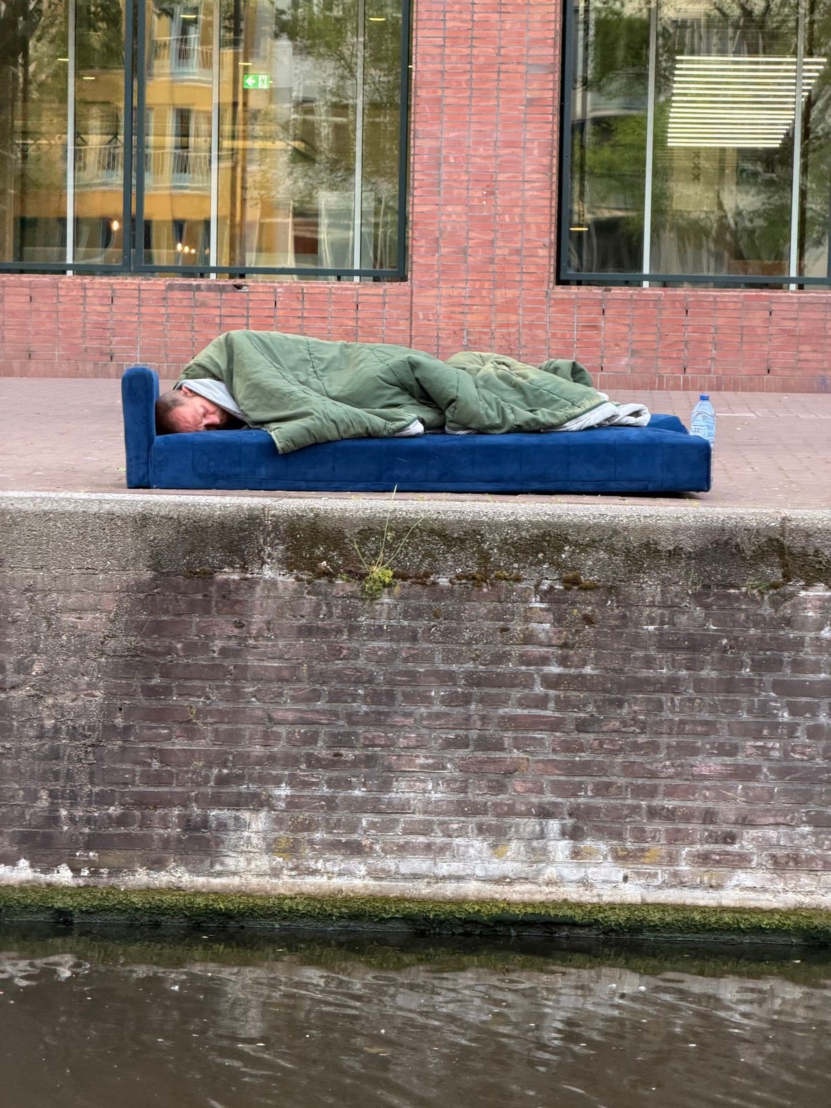 A person is seen sleeping on a blue sofa covered by a green sleeping bag, positioned on a sidewalk next to a canal