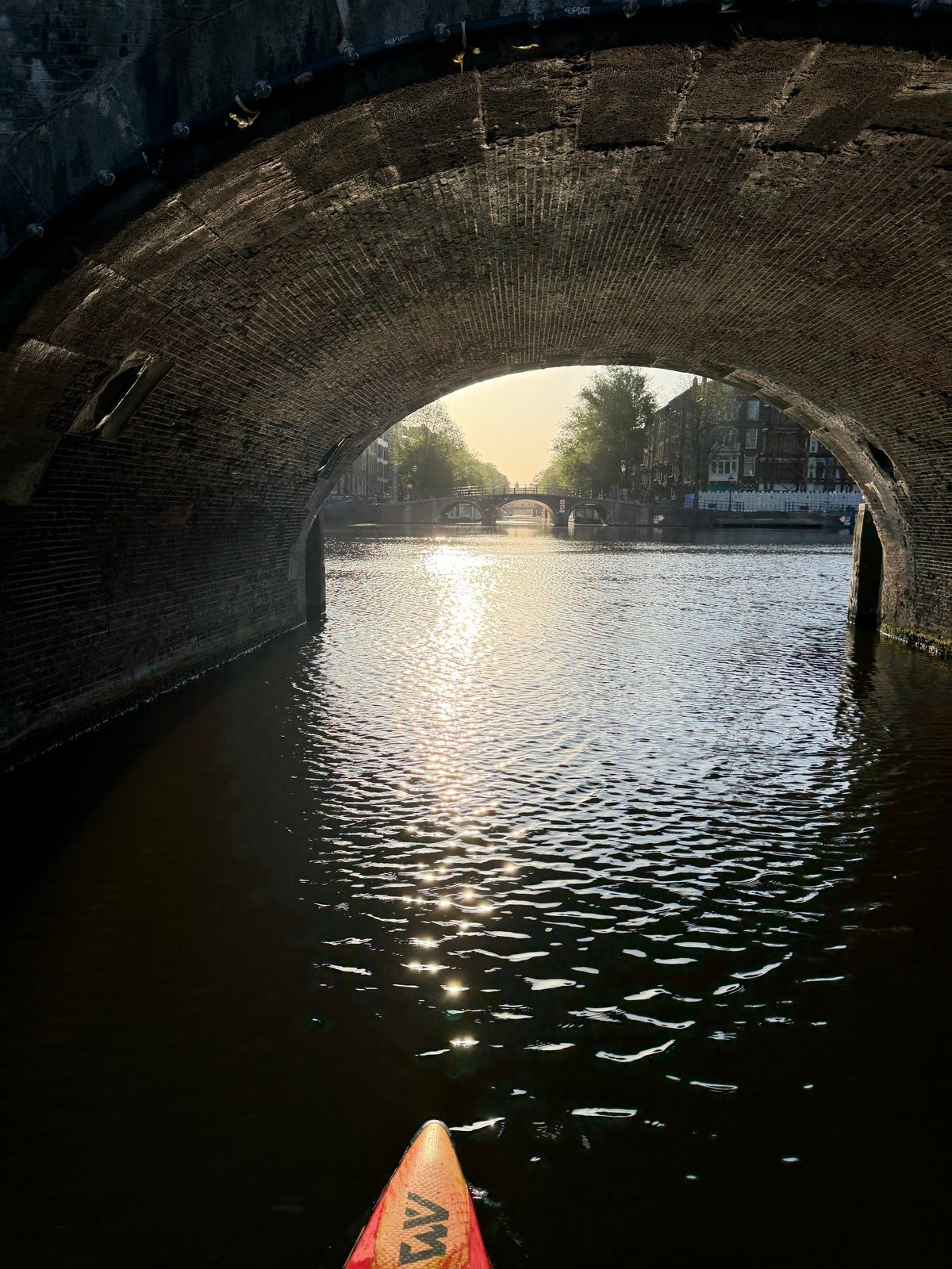 The photo captures a serene view from a paddleboard, looking out from under a dark brick bridge onto a sun-drenched canal
