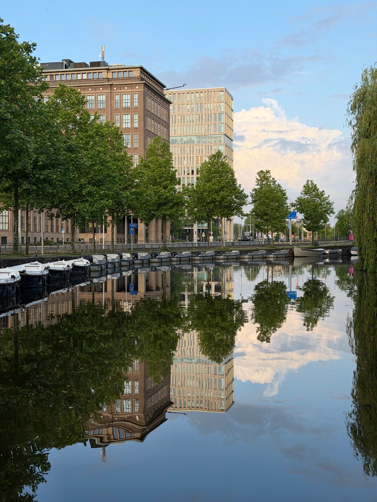 This photo captures a tranquil urban canal scene, with a row of boats docked along the water reflecting the surrounding buildings and lush green trees
