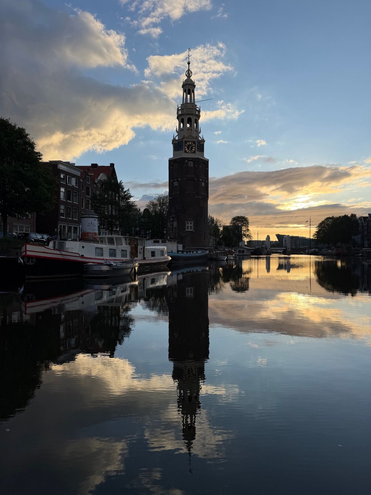 A historic clock tower, the Montelbaanstoren, stands prominently beside a tranquil canal in Amsterdam, its reflection perfectly mirrored in the calm water