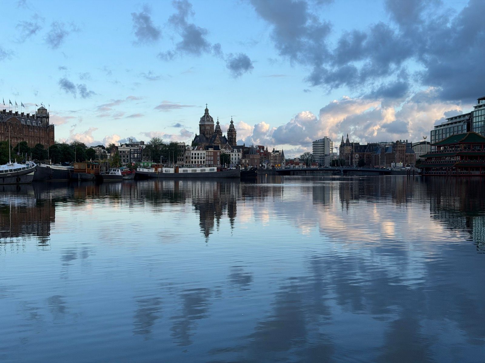 This photo captures a tranquil evening view of the Amsterdam cityscape, with the iconic Basilica of Saint Nicholas prominently reflected in the calm waters of the canal