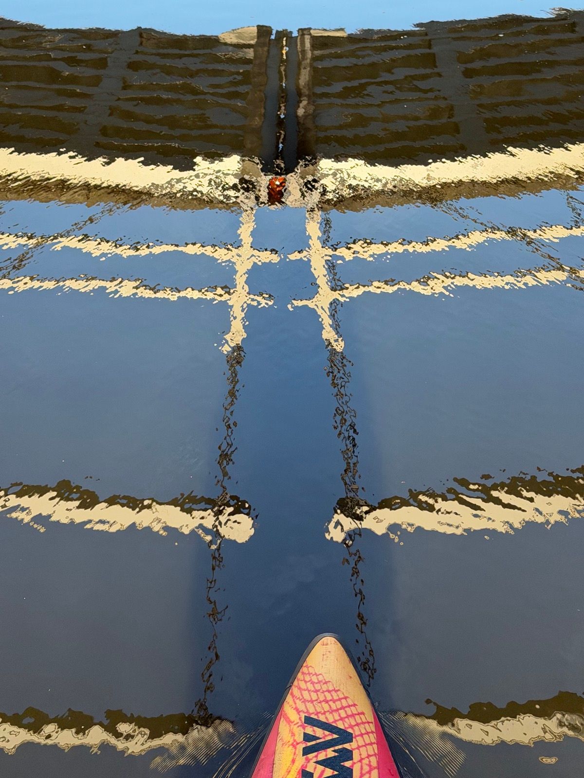 The image captures a vibrant pink and yellow paddleboard gliding across calm water, reflecting a grid-like bridge structure above
