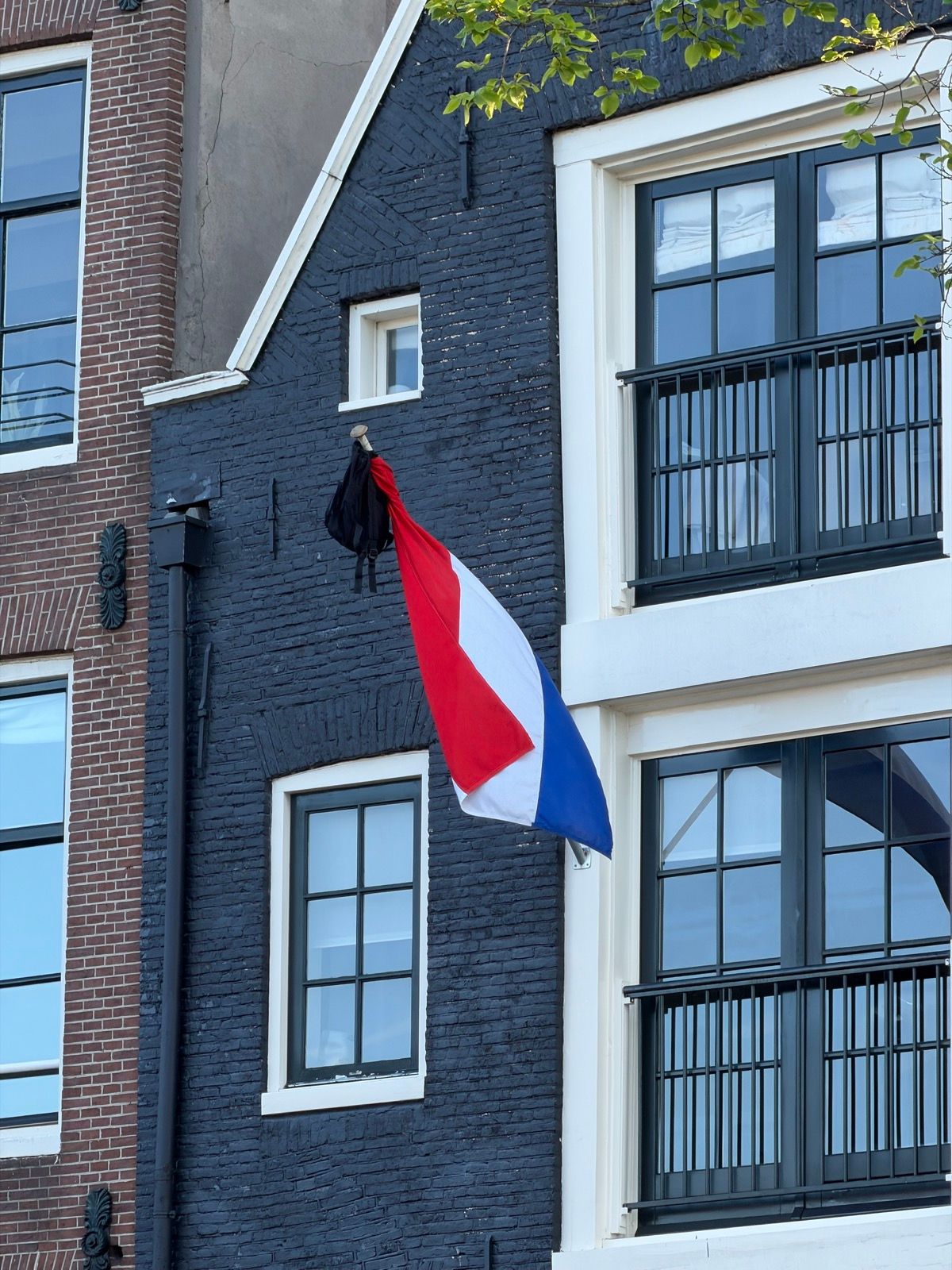 A Dutch flag, hung upside down with a black bag, is prominently displayed on the dark blue brick facade of a traditional Dutch canal house