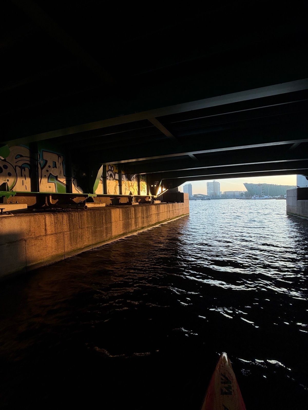 The photo captures a serene view from a paddleboard, looking out from under a dark bridge into a sunlit urban canal or river
