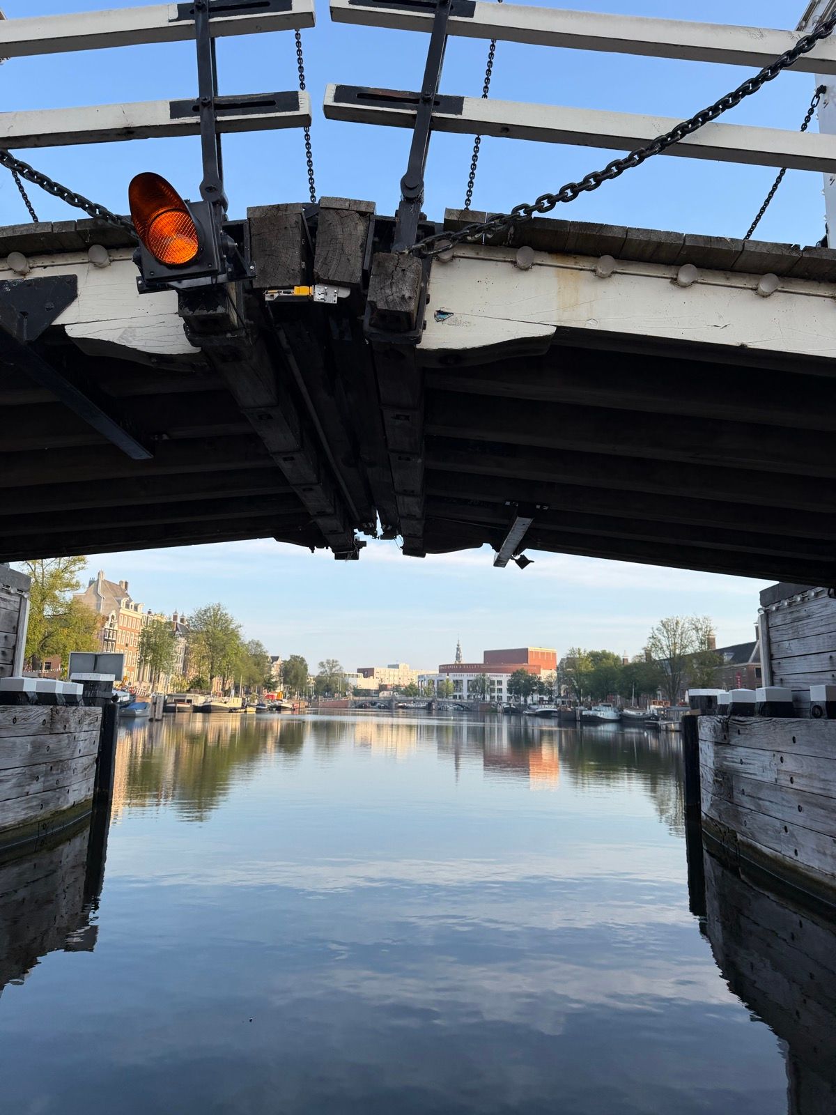 The photo captures a serene view from beneath an open drawbridge, revealing a calm canal reflecting the clear blue sky and distant city buildings
